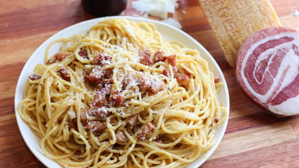 spaghetti carbonara dans bol blanc avec pancetta et bloc de parmesan sur table de bois