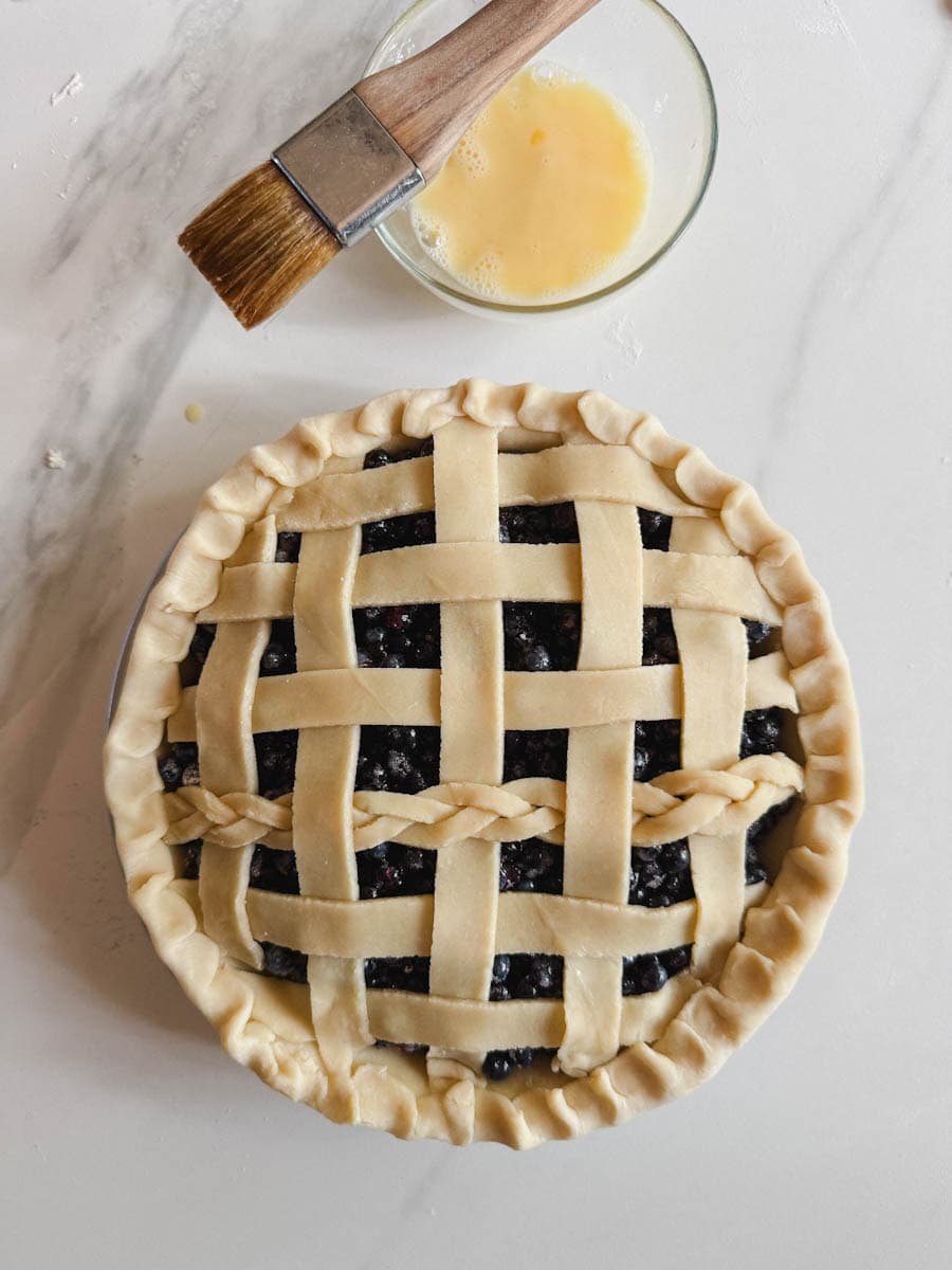 tarte aux bleuets non cuite joliment décorée avec bol de dorure à l'oeuf et pinceau