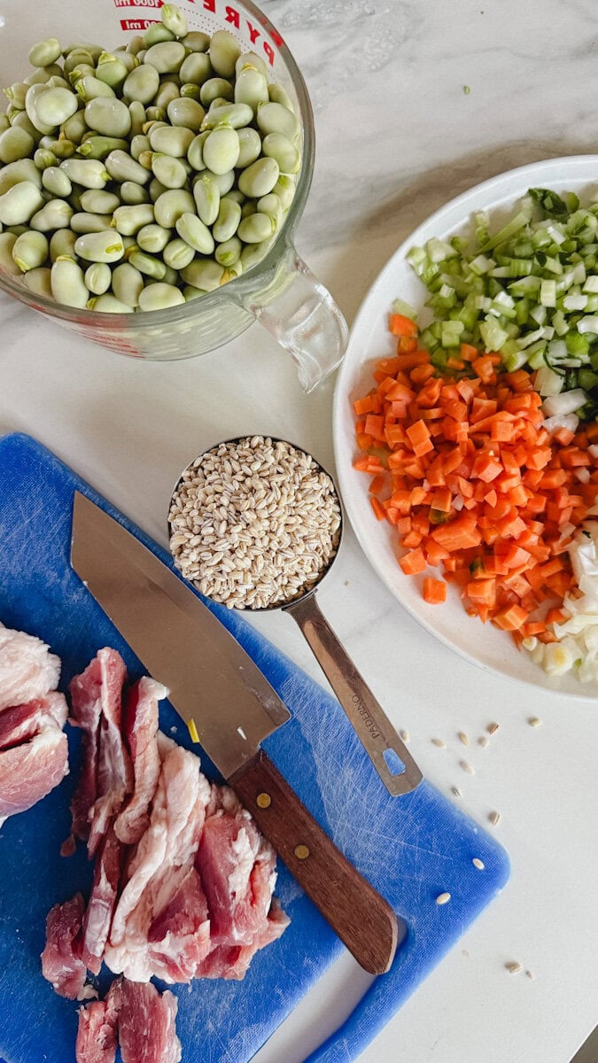 ingrédients pour soupe aux gourganes sur comptoir blanc: fèves gourganes écossées, céleri, carotte, oignon, orge, lard salé
