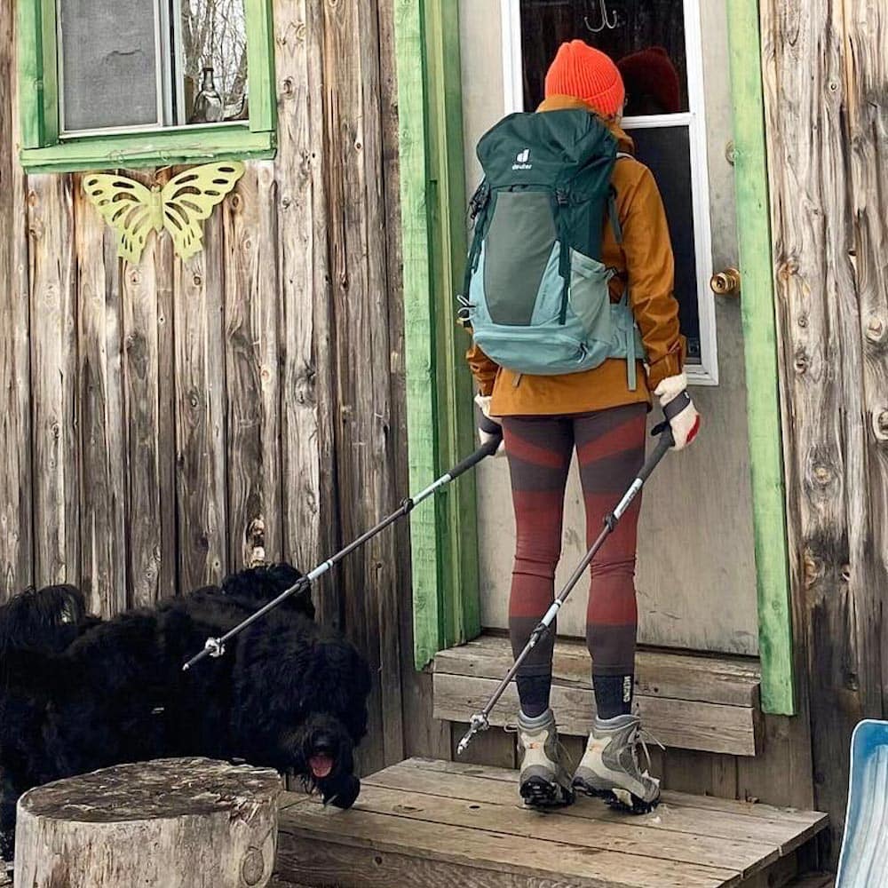 Manon lapierre de dos avec sac à dos, bâtons de marche et bottes de randonnée devant une porte de cabane