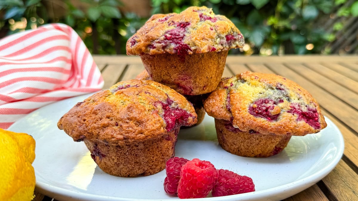 3 muffins aux framboises dans une assiette blanche avec citron et framboise dehors sur table en bois devant verdure naturelle
