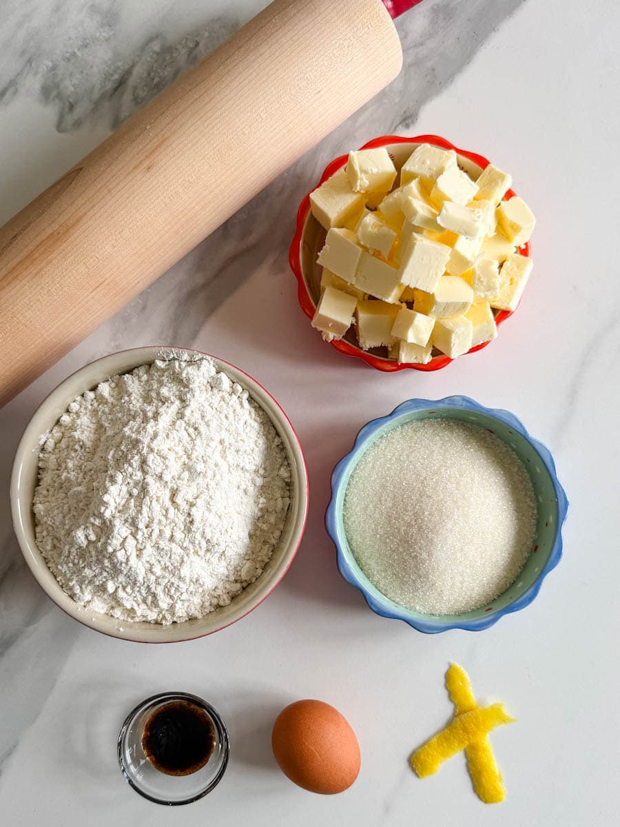 ingrédients pour biscuits emporte-pièce sur comptoir avec rouleau à pâtisserie: beurre, farine, sucre, oeuf, vanille, zeste