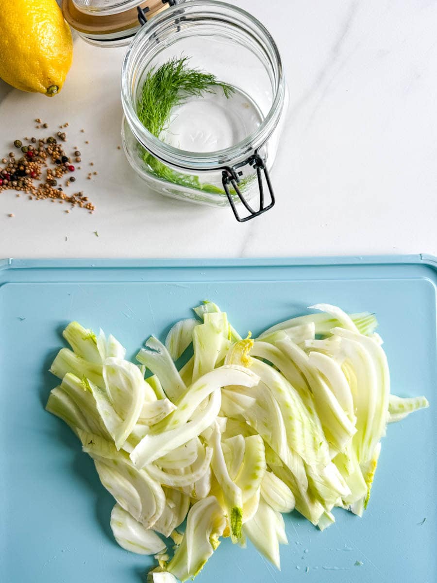 lanières de fenouil sur planche turquoise, pot à marinade vide ouvert avec citron et épices