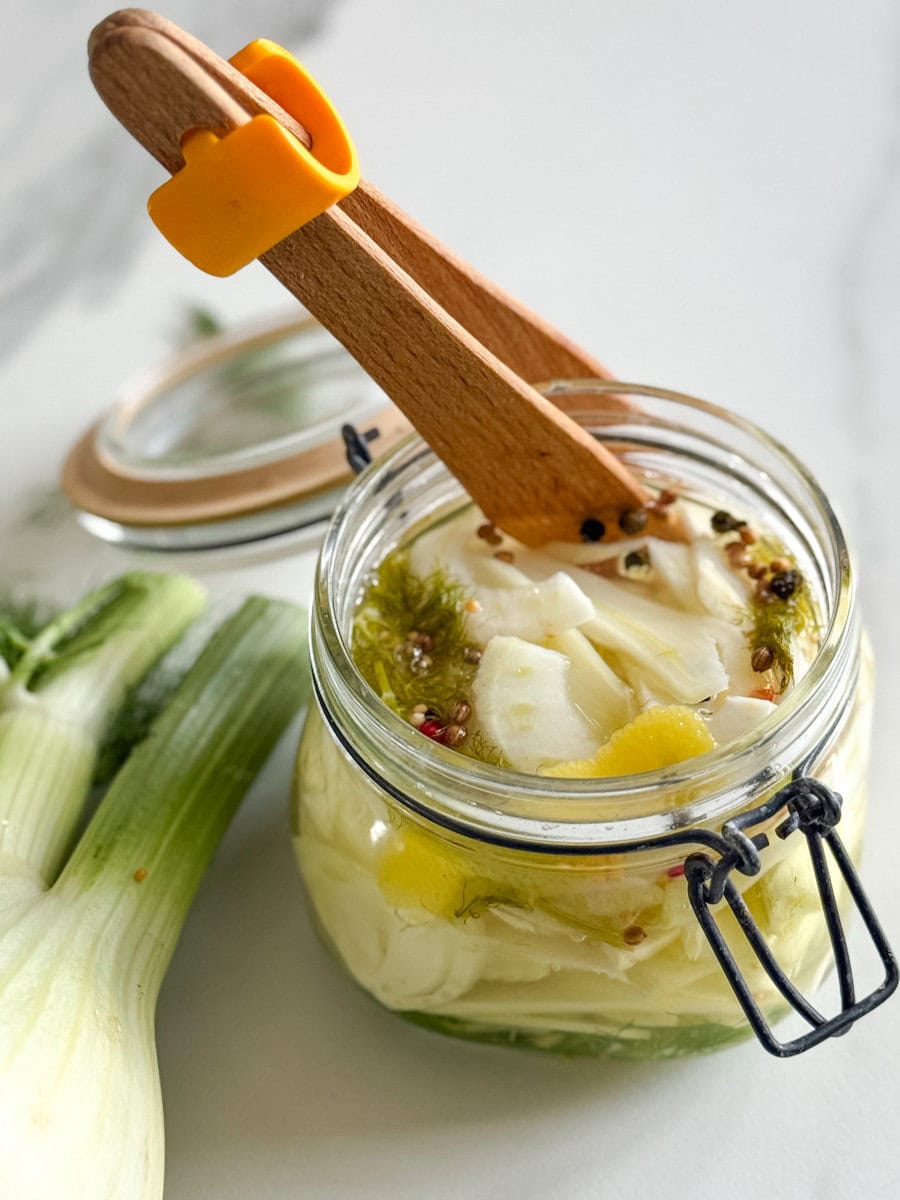 pot de marinade à couvercle avec pince en bois et bulbe de fenouil sur comptoir marbré blanc