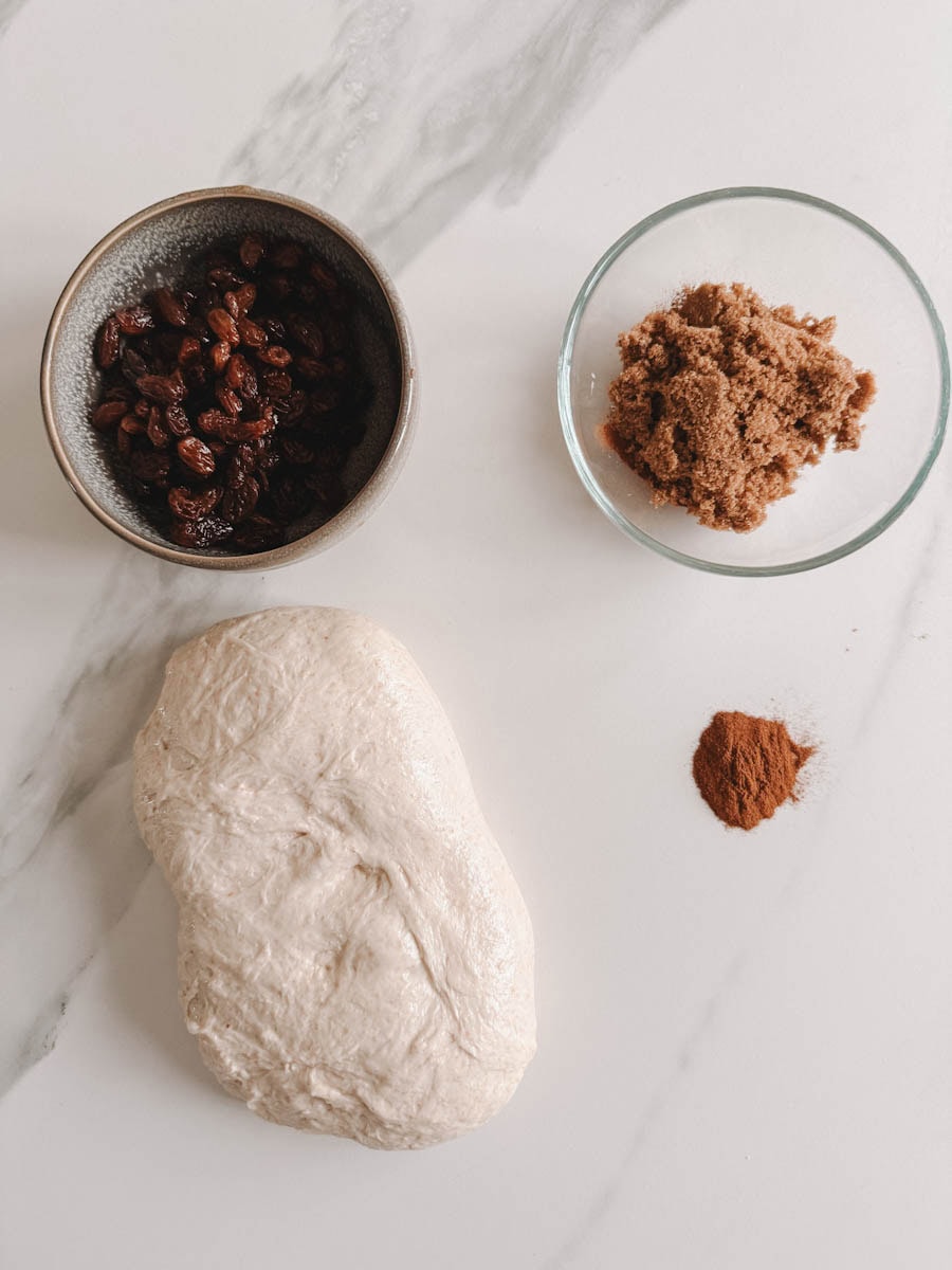 ingrédients pain aux raisins: pâte, raisins, cannelle cassonade sur comptoir marbré blanc