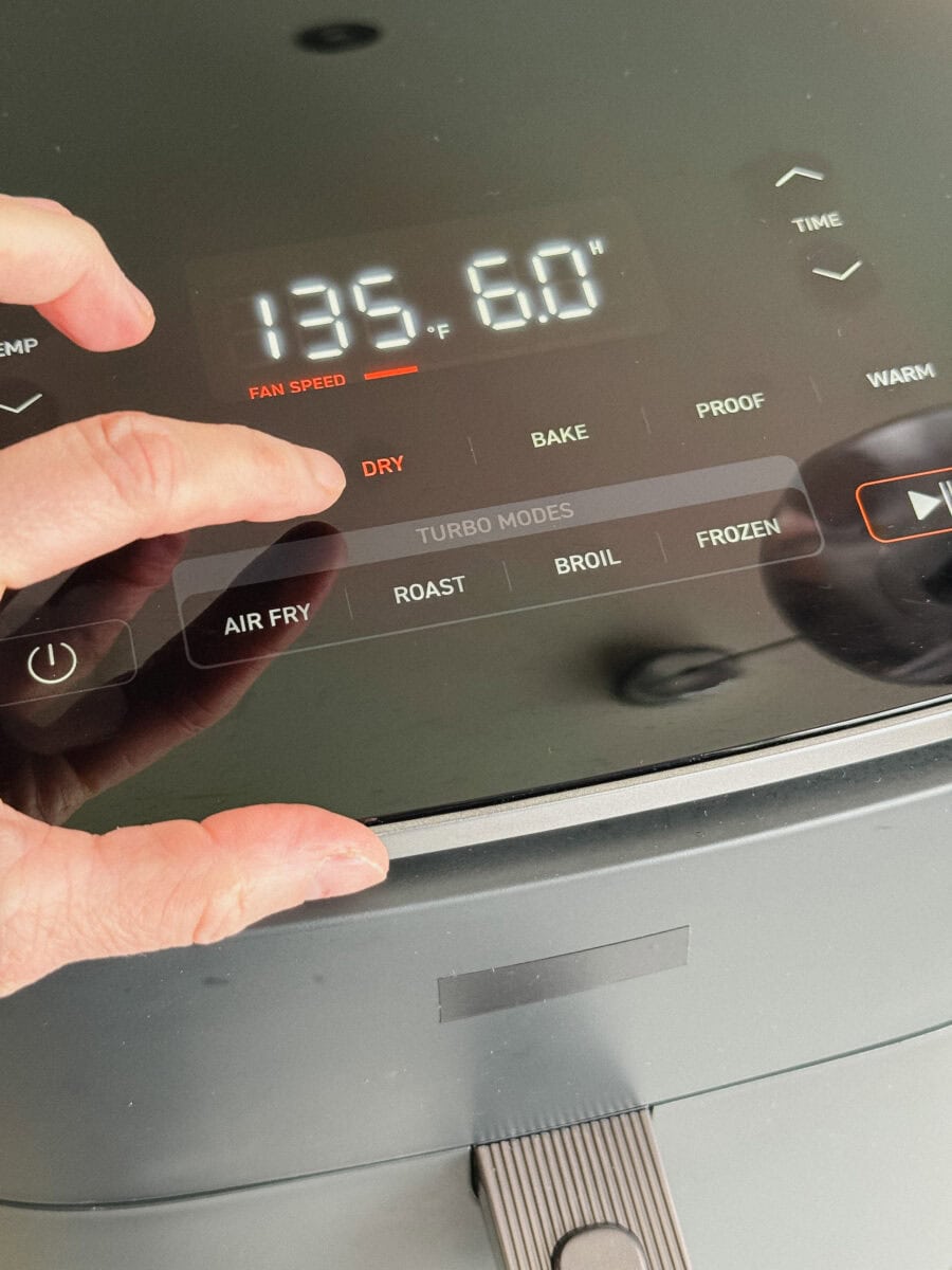 doigt qui presse le bouton Dry sur un air fryer gris foncé- on voit température et 6 heures de cuisson