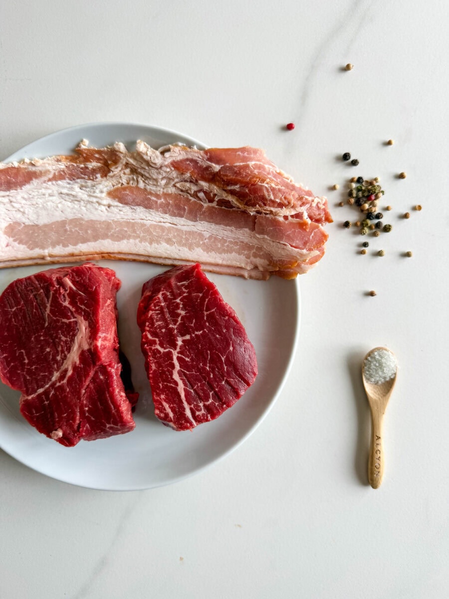 2 pièces de filet mignon de boeuf cru, tranches de bacon, sel et poivre dans assiette blance et comptoir blanc