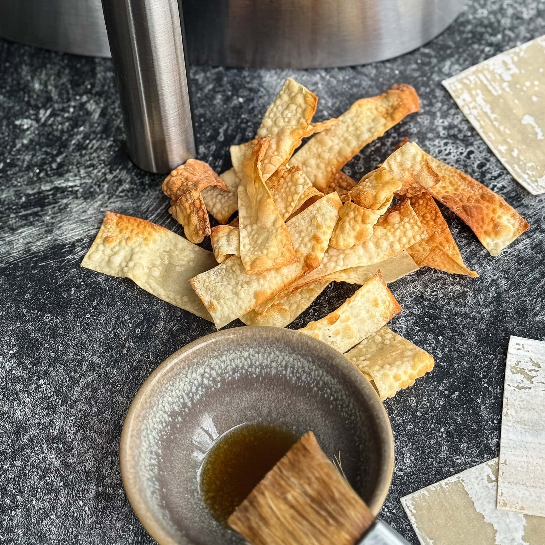 chips de wonton grillées devant panier air fryer avec petit bol d'huile grillée et pinceau à patisserie sur surface bétonnée foncée