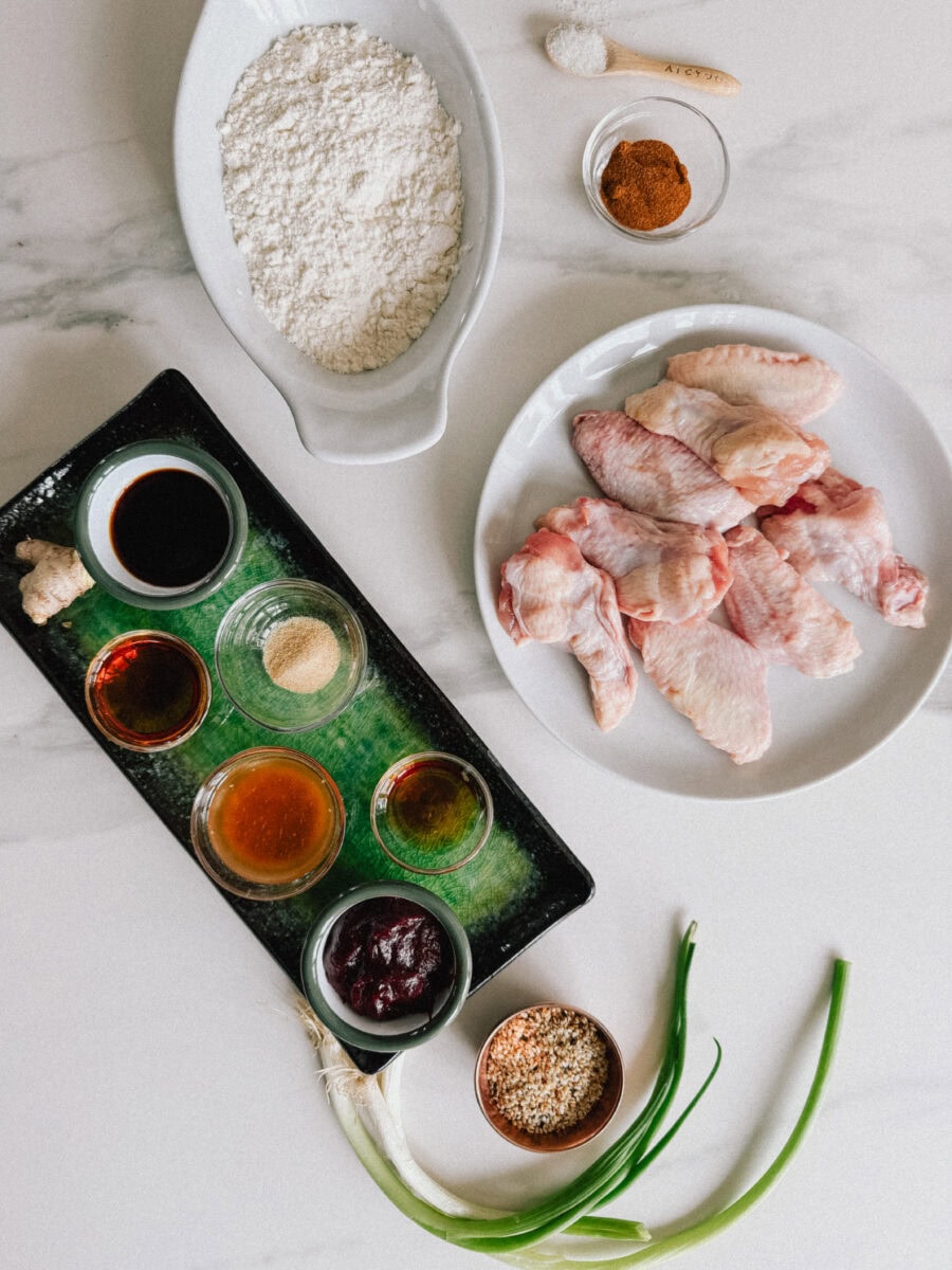ingrédients sur comptoir blanc: ailes de poulet, bol de farine, ingrédients de la sauce, oignon vert, graines de sésame