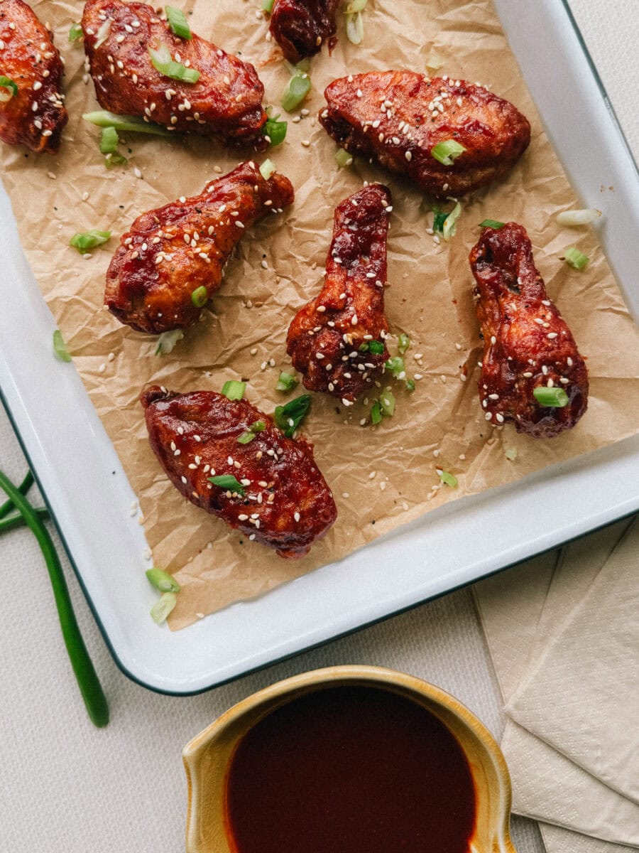Ailes de poulet glacées en sauce rouge sur plateau couvert de papier parchemin avec sésame et oignons verts et petit pot de sauce gochujang à côté