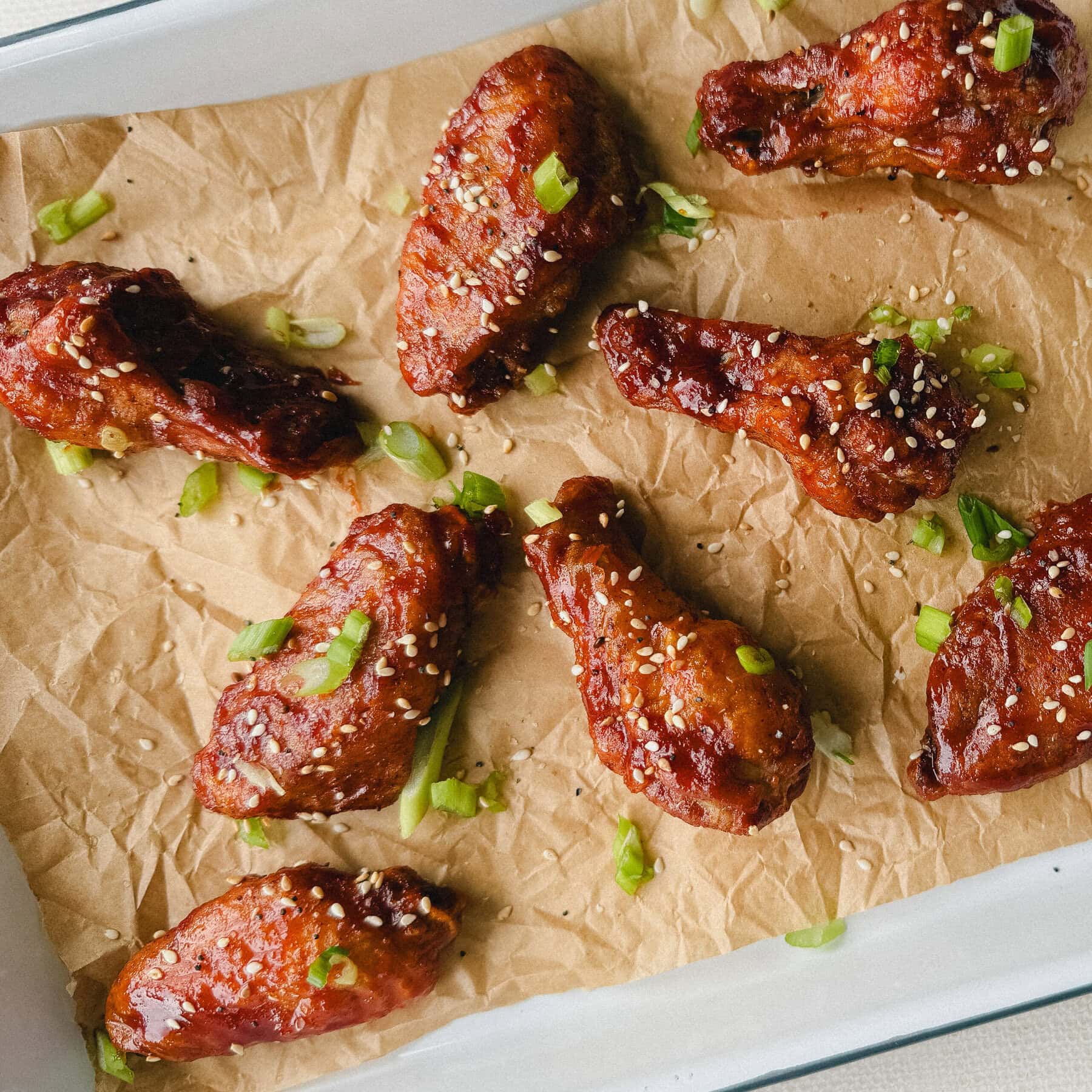 ailes de poulet glacées de sauce rouge gochujang et garnies de graines de sésame et oignon vert sur papier parchemiin brun dans un plateau