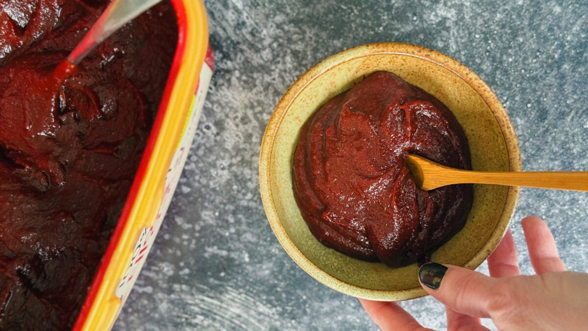 pâte gochujang dans petit bol avec cuillère en bois tenu par une main devant gros pot de gochujang