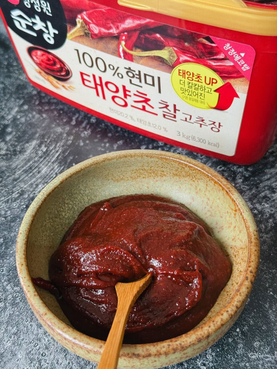 pâte gochujang dans un petit col avec cuillère en bois devant gros pot de gochujang écrit en coréen