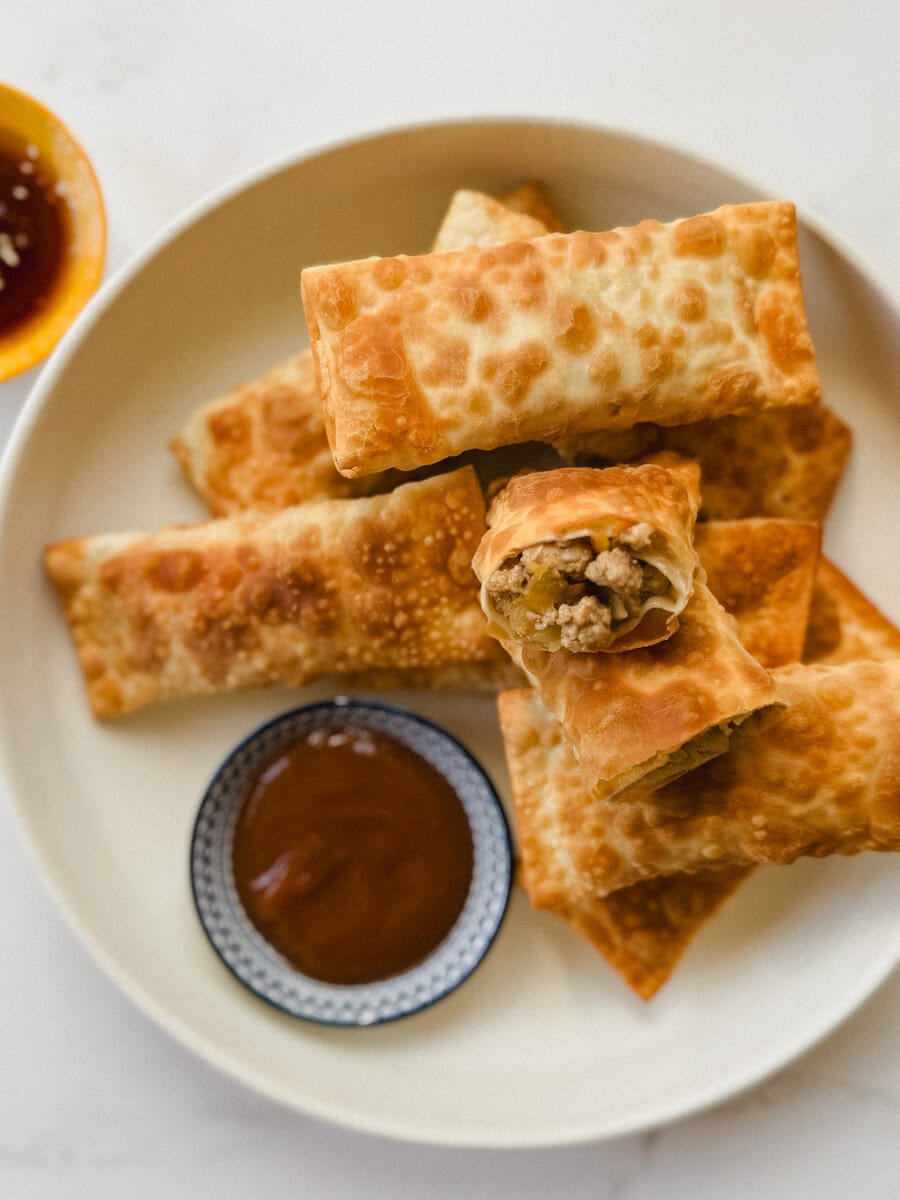 plusieurs egg rolls dans une assiette ivoire avec petit bol de sauce sur comptoir blanc