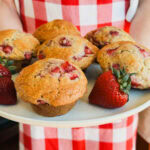 personne en tablier à carreaux rouge et blanc qui tient assiette de 6 gros muffins aux fraises