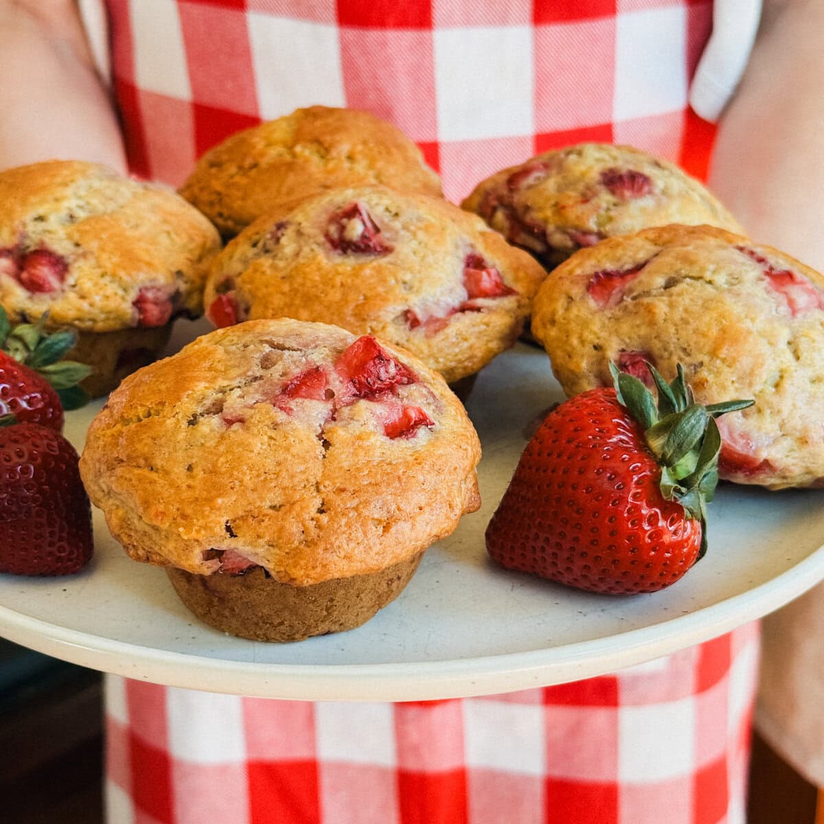 personne en tablier à carreaux rouge et blanc qui tient assiette de 6 gros muffins aux fraises