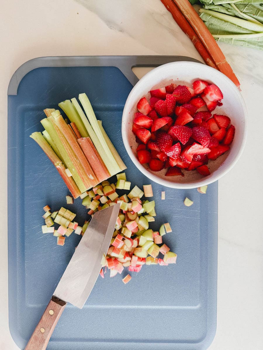 couteau, rhubarbe coupée et tiges de rhubarbe sur planche à découper bleu et bol de fraises en morceau