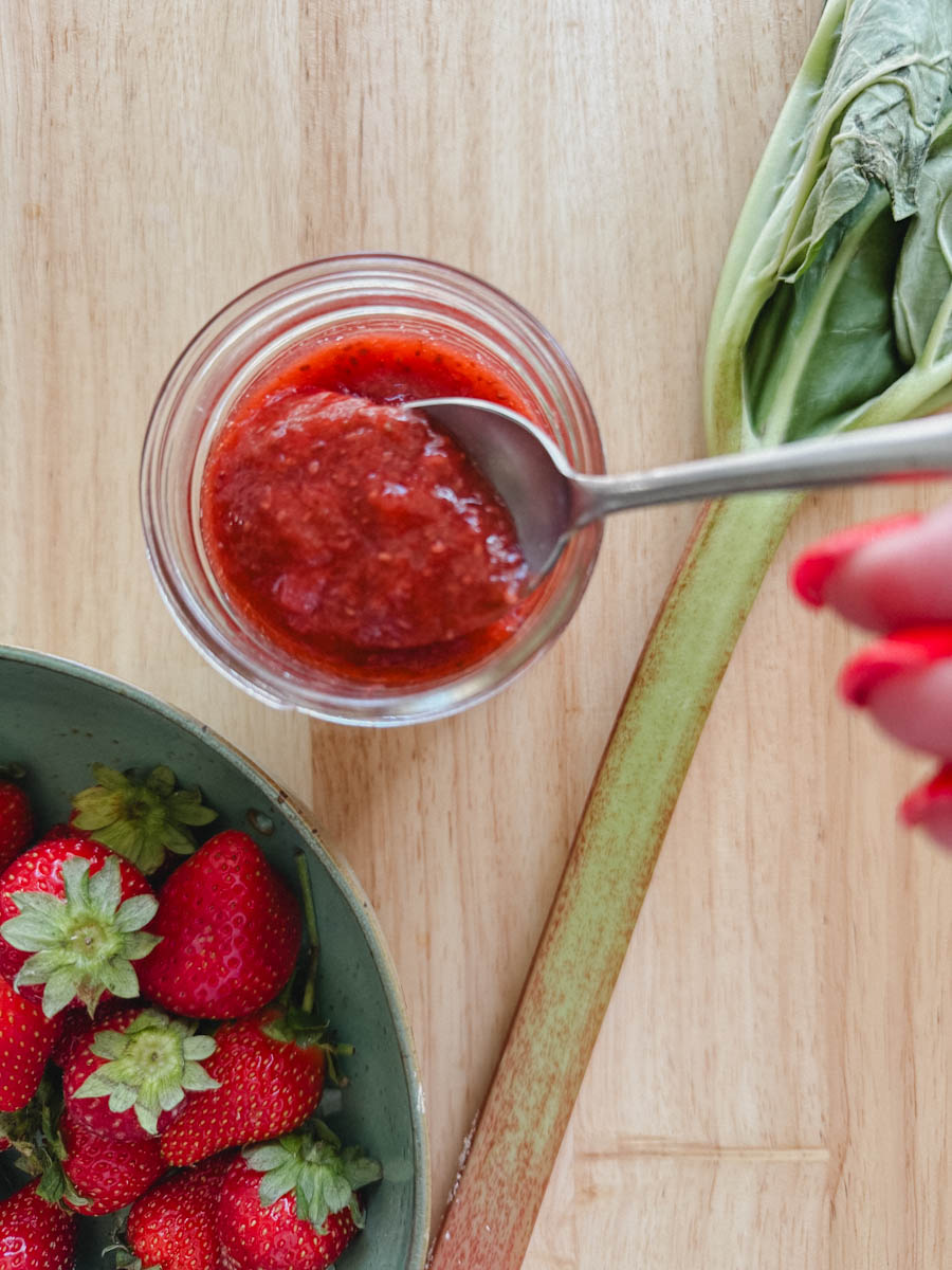 main qui tient une cuillère dans un pot de confiture fraise rhubarbe avec branche de rhubarbe et fraises fraîches à côté