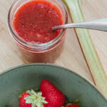 pot de confiture fraise rhubarbe avec cuillère à l'intérieur, tige de rhubarbe et bol de fraises à côté