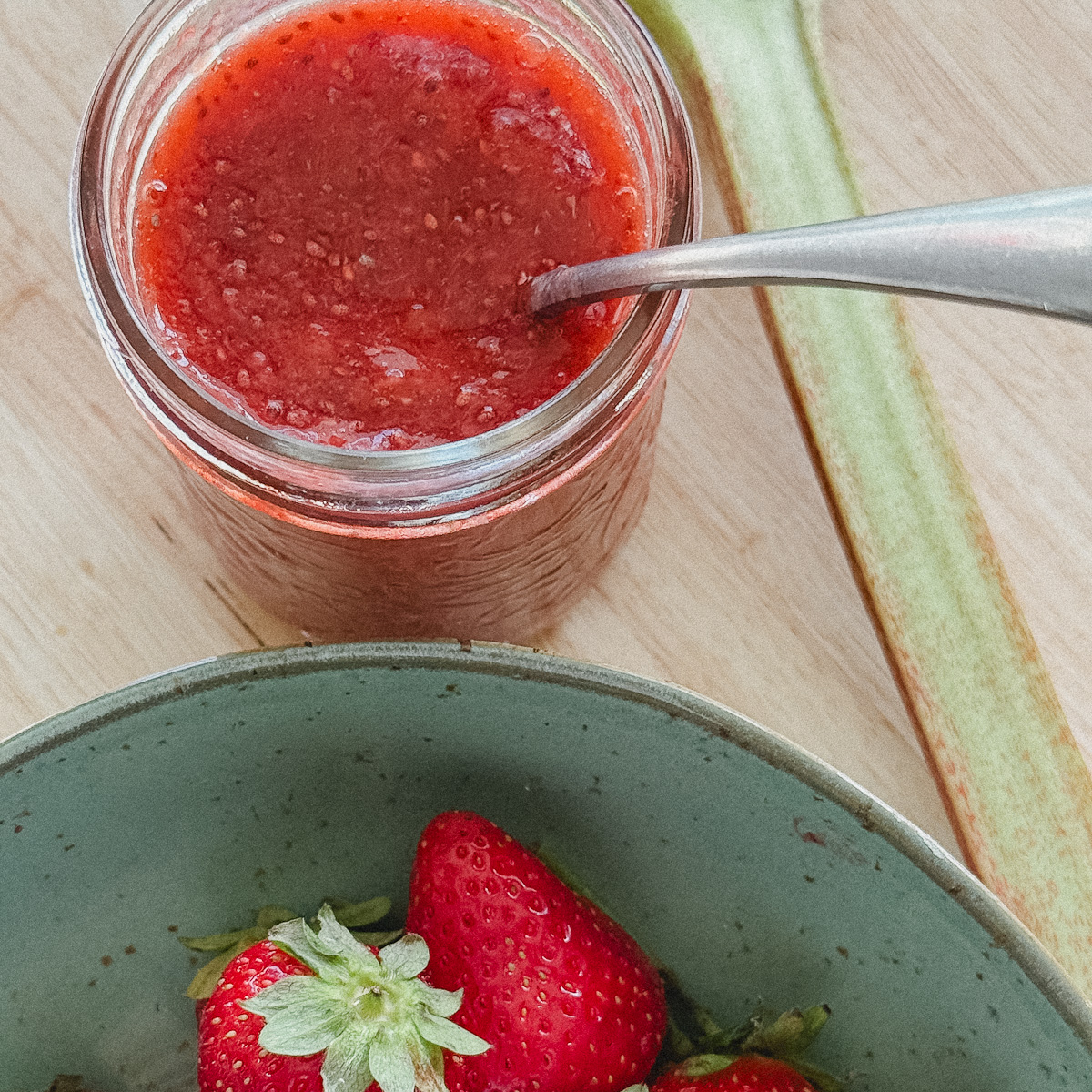 pot de confiture fraise rhubarbe avec cuillère à l'intérieur, tige de rhubarbe et bol de fraises à côté