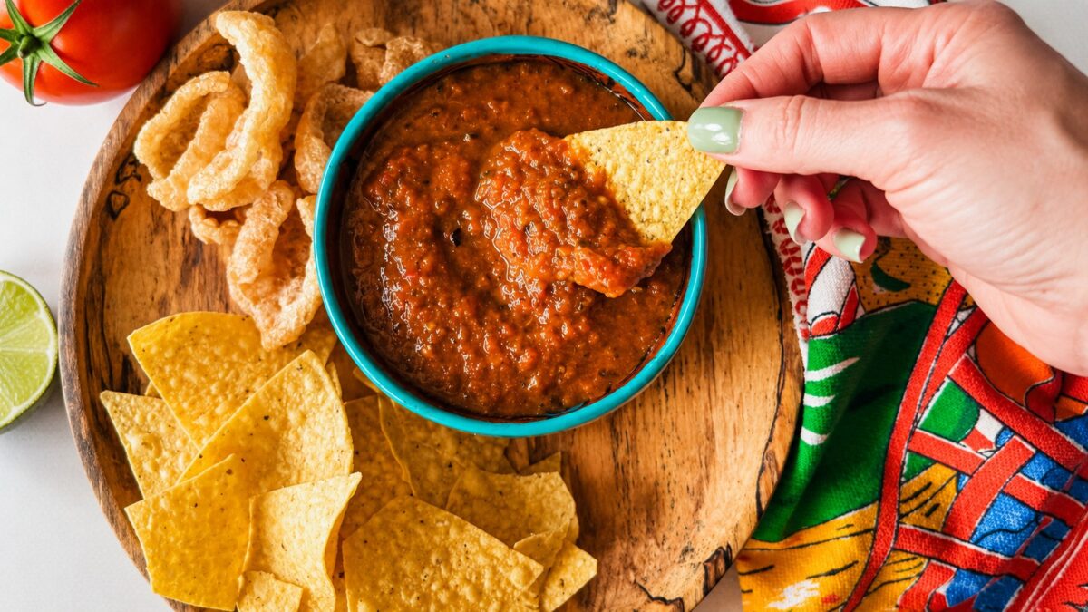 main tenant une chips de maïs qui trempe dans un bol de salsa mexicaine dans une assiette en bois