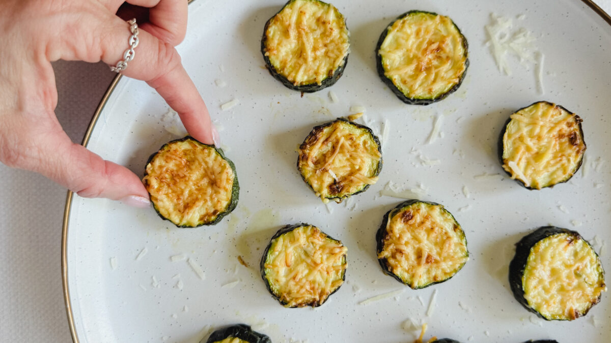 main qui tient une rondelle de courgette gratinée dans une assiette avec plusieurs autres