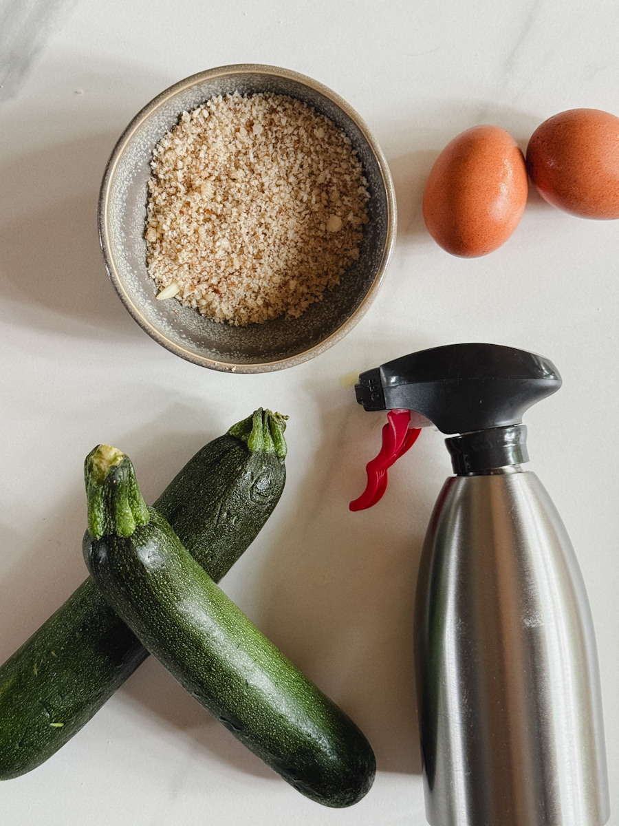 ingrédients sur comptoir blanc marbré: 2 courgettes, bouteille de vaporisateur, bol de chapelure et 2 oeufs