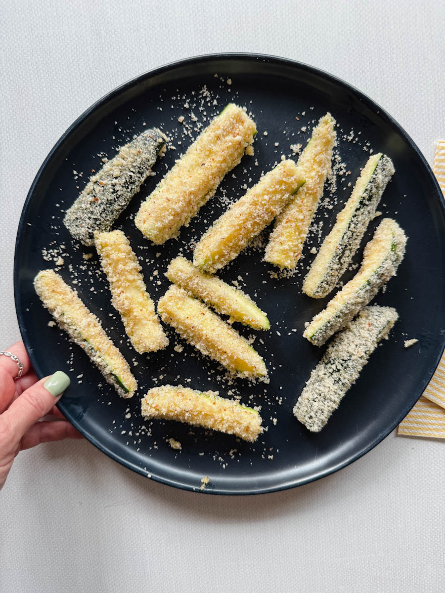 main qui tient une assiette bleu foncée de frites de courgettes panées non cuites 