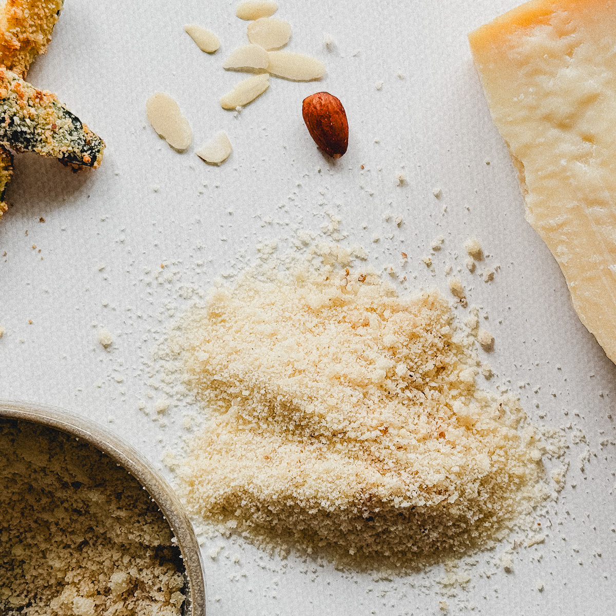 chapelure amande et parmesan sur comptoir blanc avec amandes, bloc de parmesan et bol en poterie beige