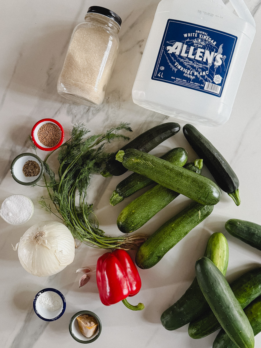 ingrédients pour relish maison sur comptoir blanc marbré: concombres, courgettes, oignon, ail, épices, vinaigre en bidon