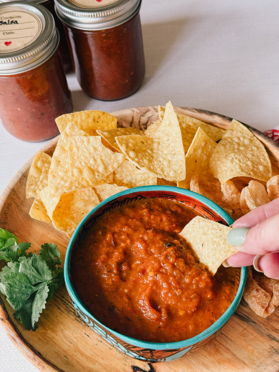 main qui tient une chips de maïs dans un bol de salsa, pots de conserve de salsa maison en arrière plan