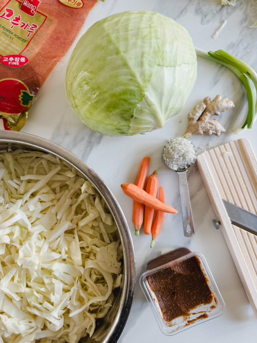 ingrédients pour kimchi sur comptoir blanc marbré: bol de chou, chuu entier, carottes, oignon vert, gingembre, gochugary et pâte miso rouge