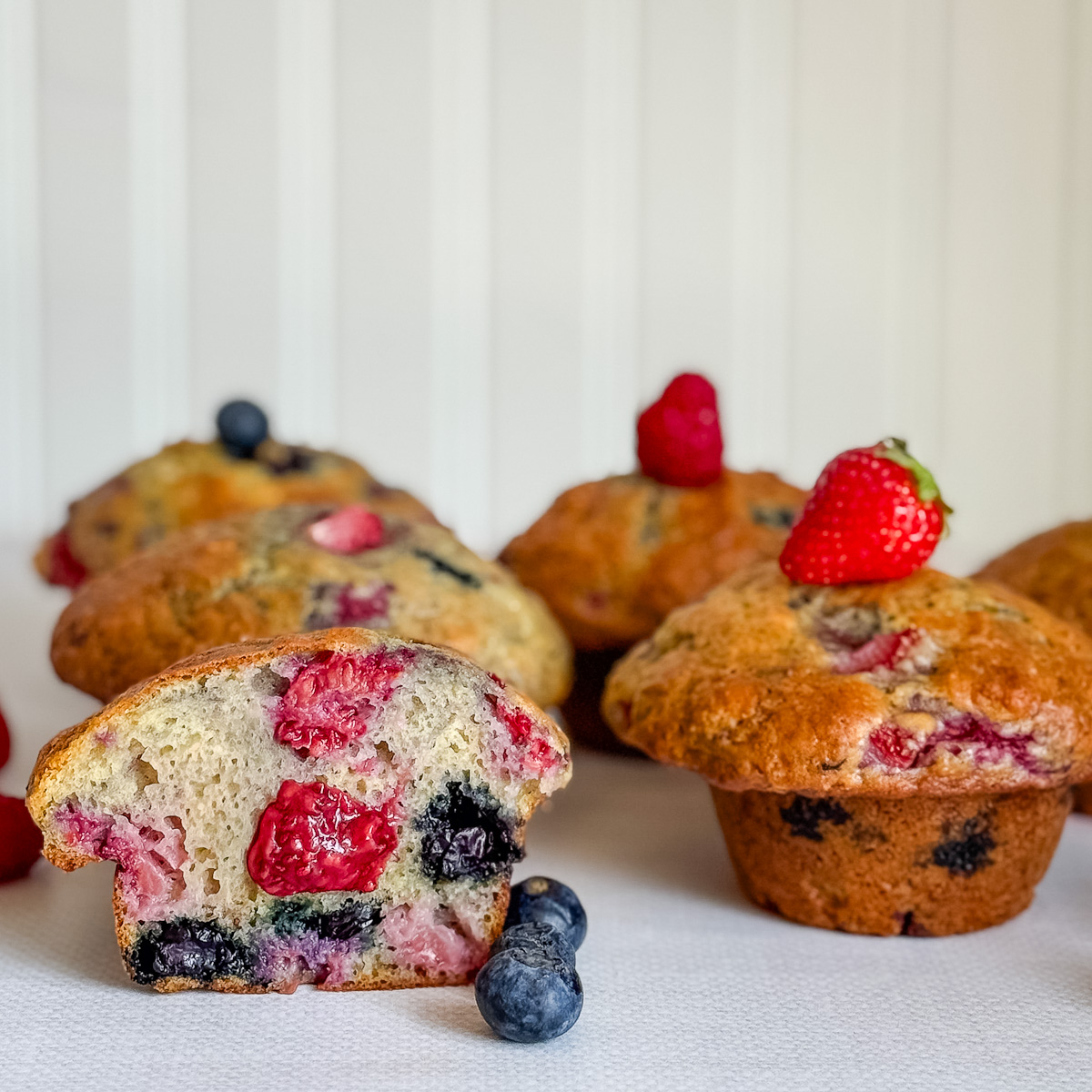 4 gros muffins avec un coupé au centre avec confiture visible et petits fruits, bleuets, fraises, framboises sur table blanche