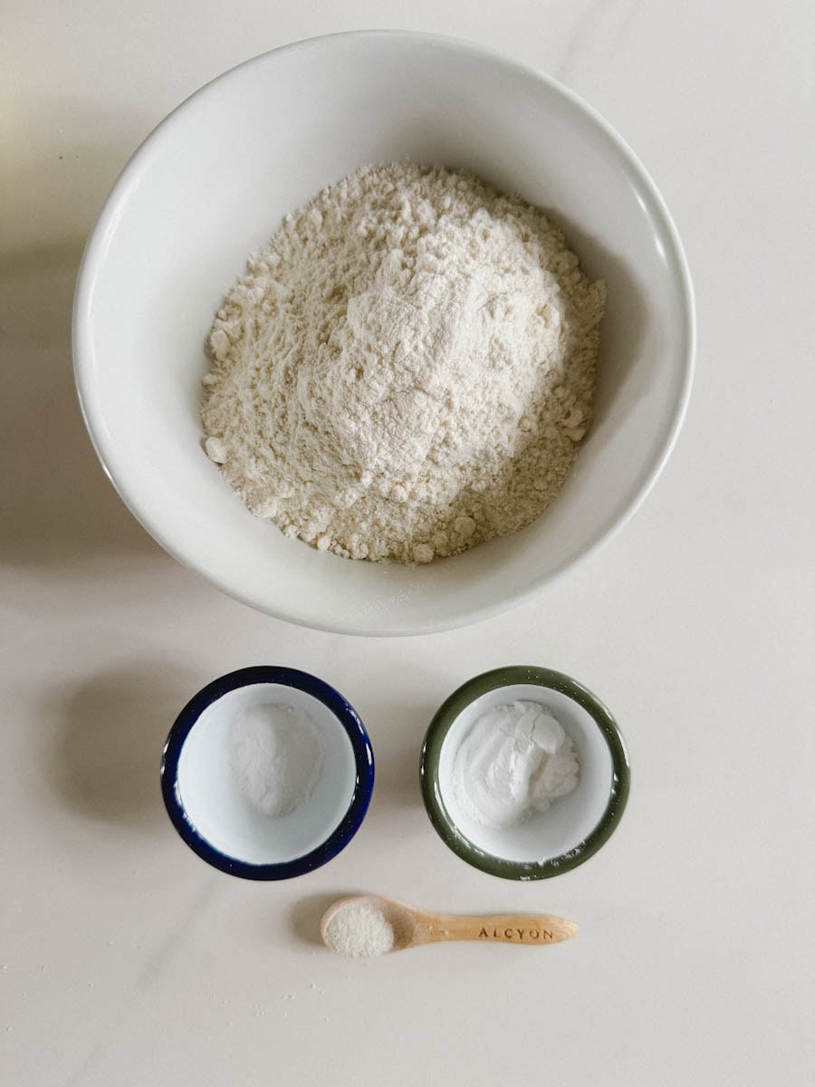 bol de farine, petits bols de poudre à pâte et bicarbonate et cuillère avec du sel sur comptoir blanc marbré