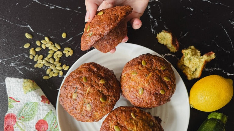 main qui tient un gros muffin aux courgettes au dessus d'une assiette avec 3 muffins, citron, zucchini et graines de citrouille sur comptoir