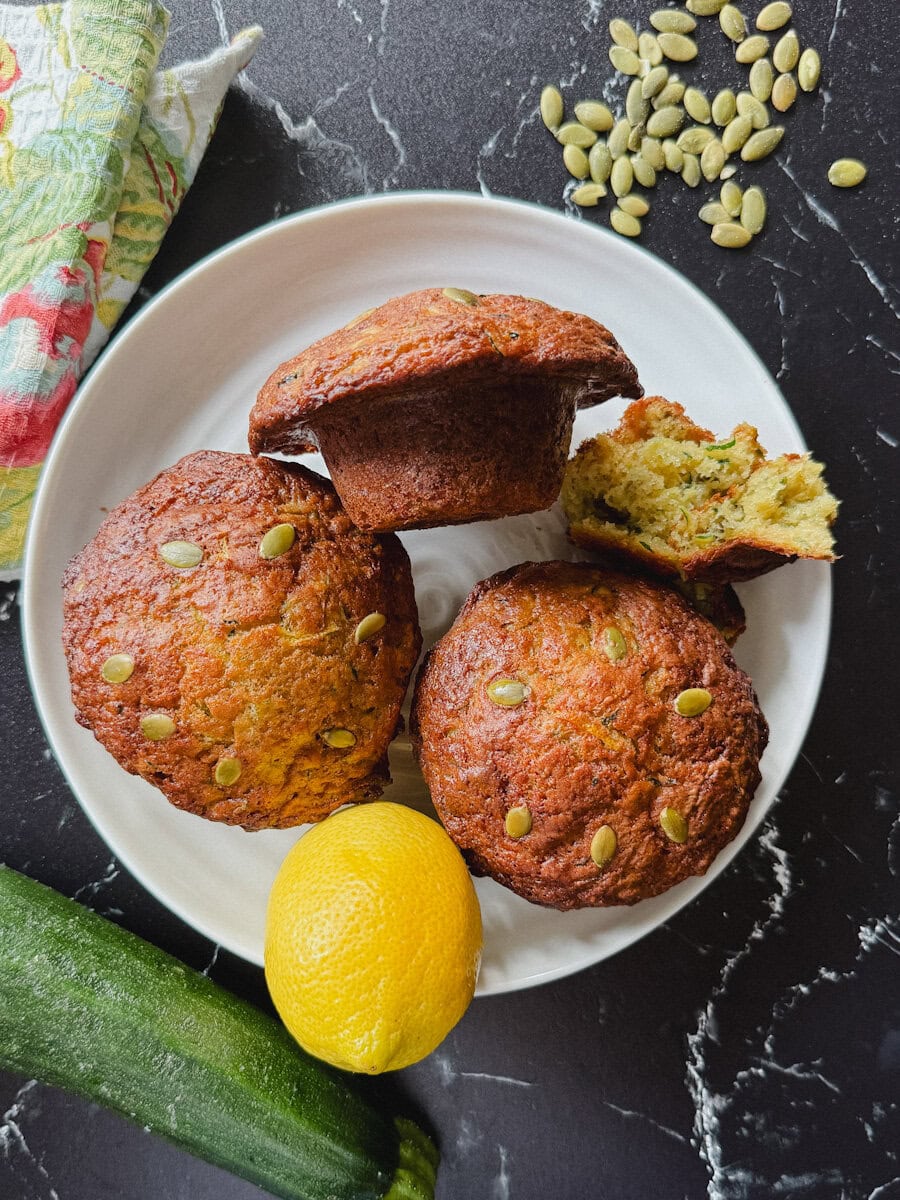 trois muffins aux courgettes bien grillés dans une assiette blanche sur comptoir noir marbré avec citron, bout de courgette et graine de citrouille alentour