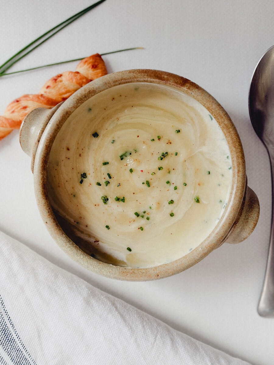 bol beige en poterie contenant crème de poireaux avec cuillère, paille au fromage, quelques brins de ciboulette, à côté