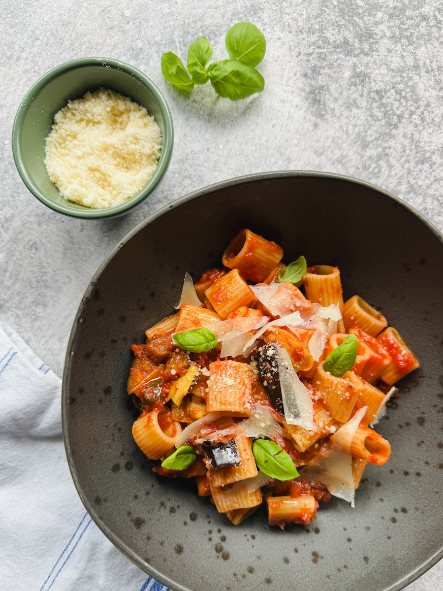 bol de pâtes sauce rouge aubergine et artichauts avec bol de fromage râpé et basilic sur fond béton