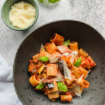 pâtes aubergines et artichauts dans un bol noir tacheté avec petit bol de pecorino râpé à côté et basilic frais sur comptoir style béton gris pâle