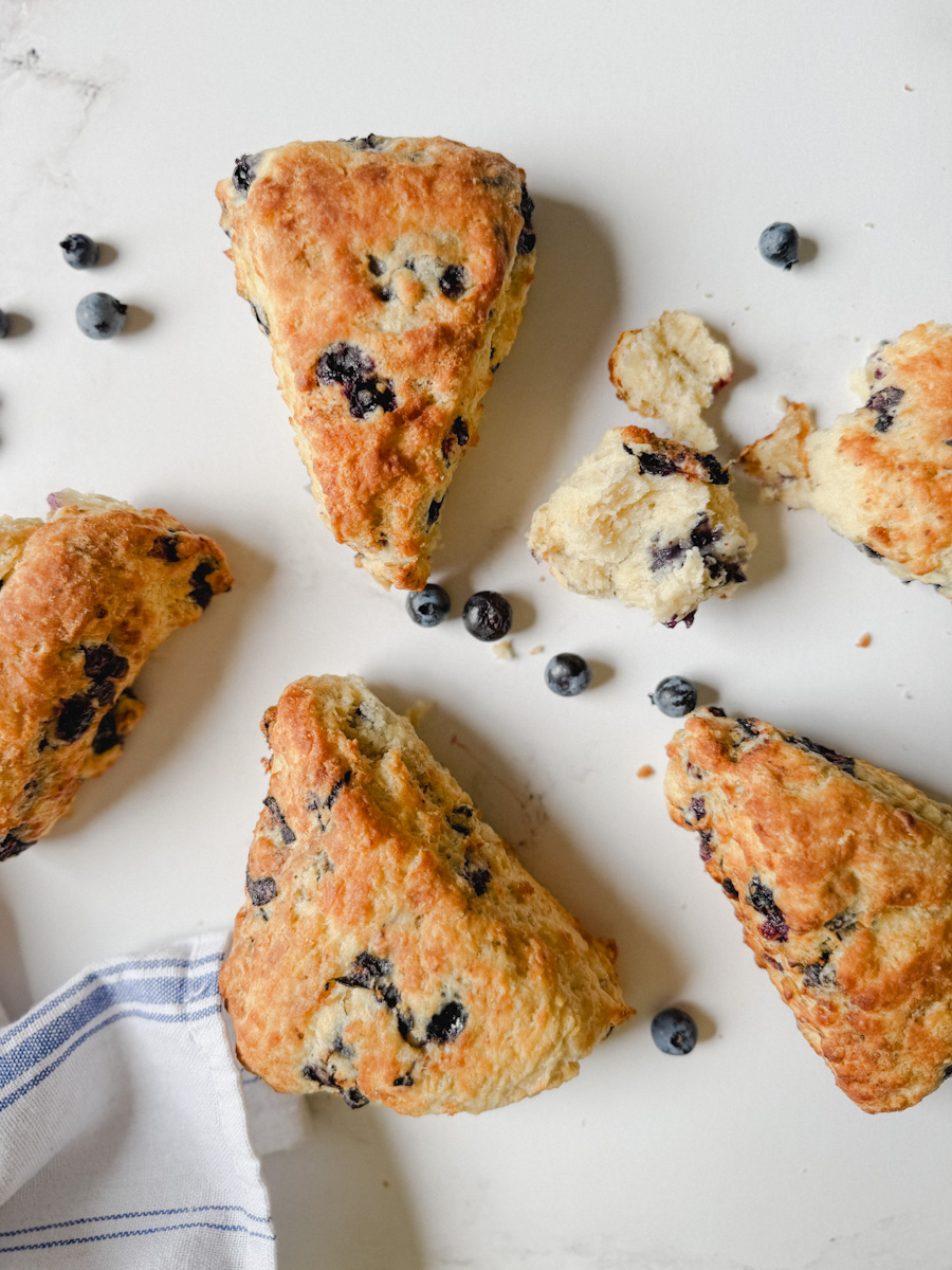 4 scones aux bleuets sur comptoir blanc avec bleuets alentour et morceau de scone ouvert