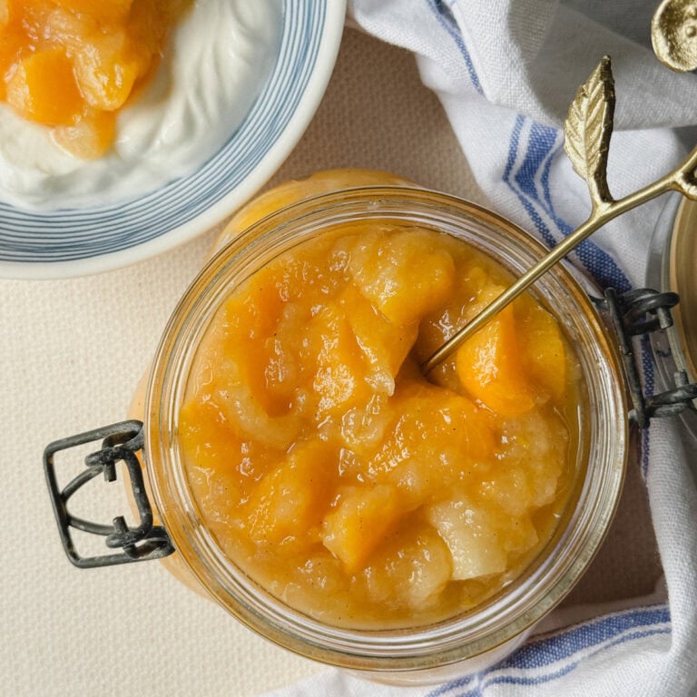 pot de compote avec dés de pomme et pêche et cuillère à l'intérieur, bol de yogourt garni de compote en arriere plan