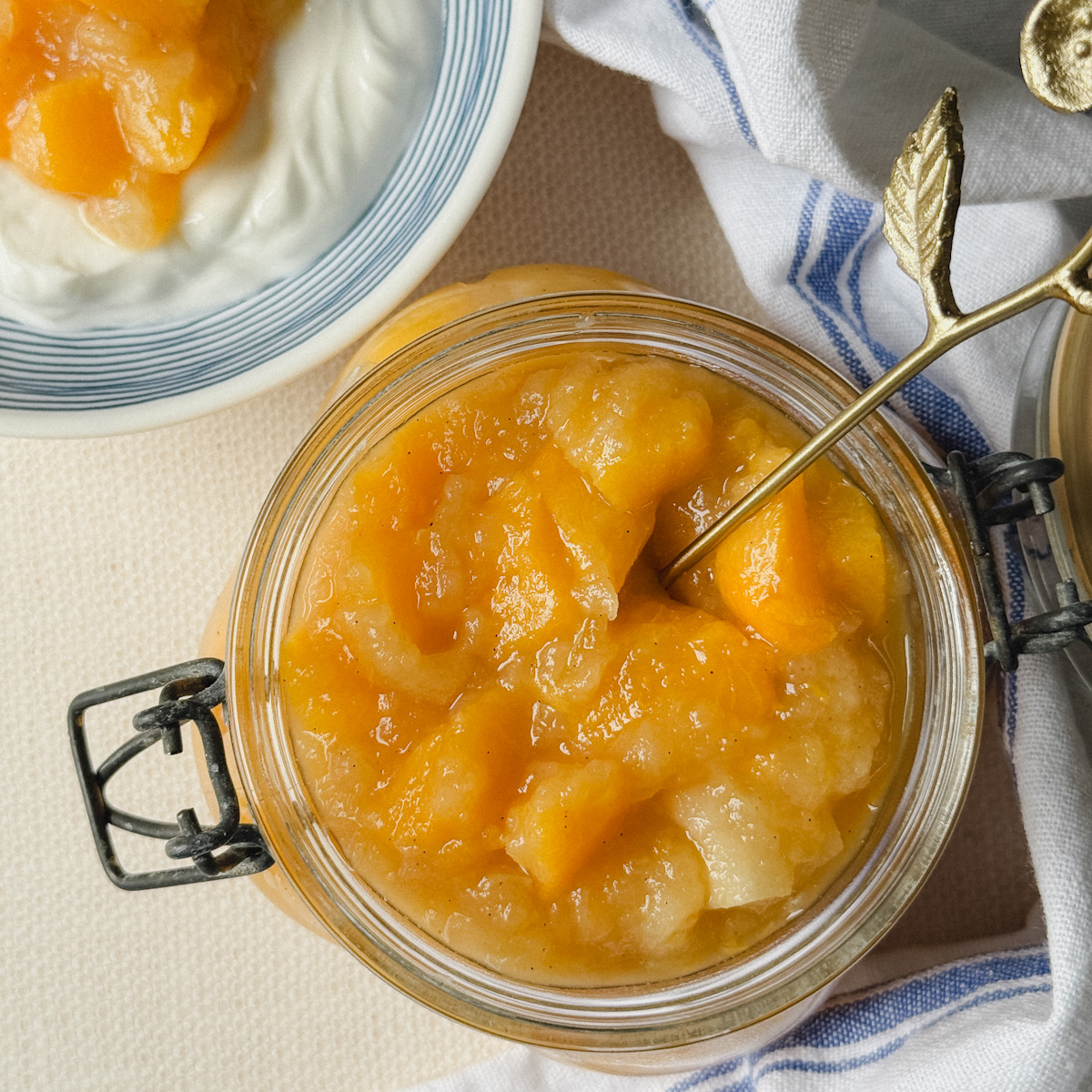 pot de compote avec dés de pomme et pêche et cuillère à l'intérieur, bol de yogourt garni de compote en arriere plan