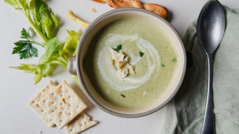 crème de céleri dans un bol en poterie sur table avec cuillère, linge de table, biscuits soda, paille au fromage et feuilles de céleri alentour