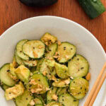 salade de concombre et avocat garnie d'arachides dans un bol blanc avec chopsticks, avocat et 1 concombre en arriere plan