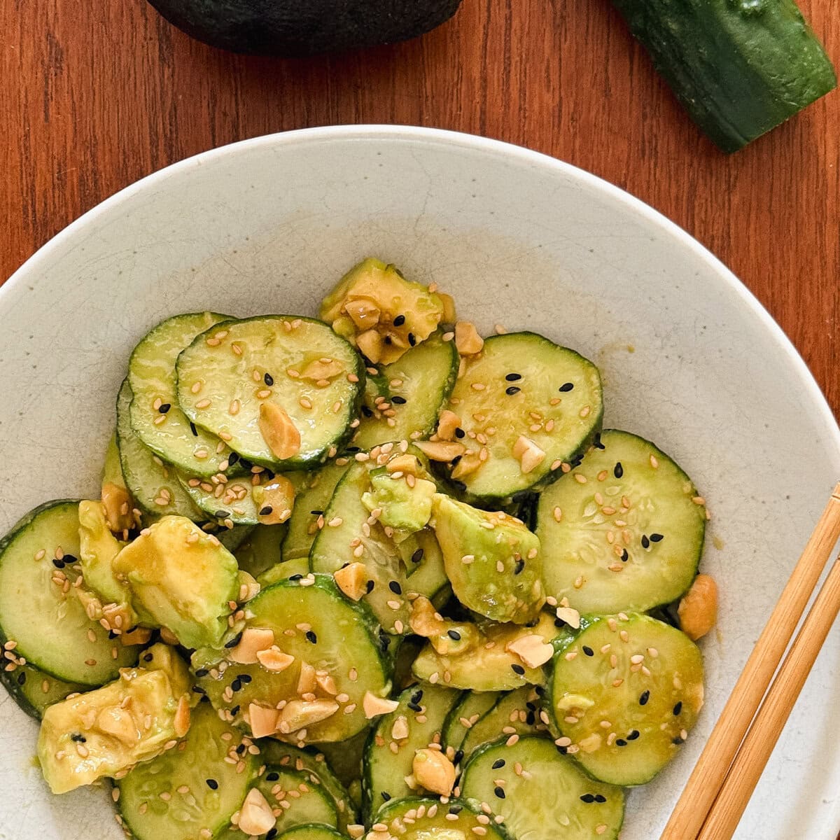salade de concombre et avocat garnie d'arachides dans un bol blanc avec chopsticks, avocat et 1 concombre en arriere plan