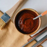 sauce bbq maison pour côtes levées et poulet dans bol avec cuillère, pinceau à pâtisserie et pinces bbq à côté