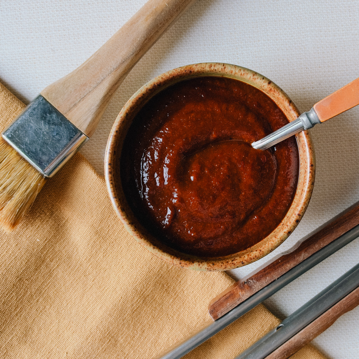 sauce bbq maison pour côtes levées et poulet dans bol avec cuillère, pinceau à pâtisserie et pinces bbq à côté