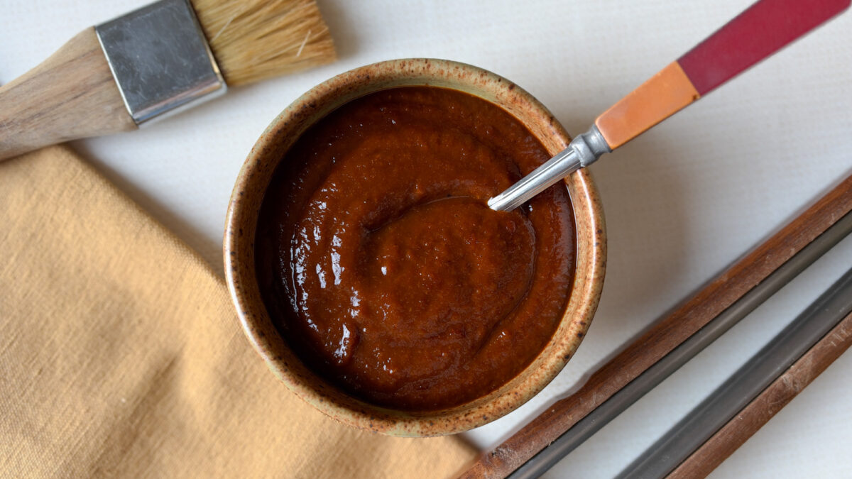 sauce bbq maison dans un bol de poterie avec cuillère, à côté de pinceau à bbq et pinces à bbq
