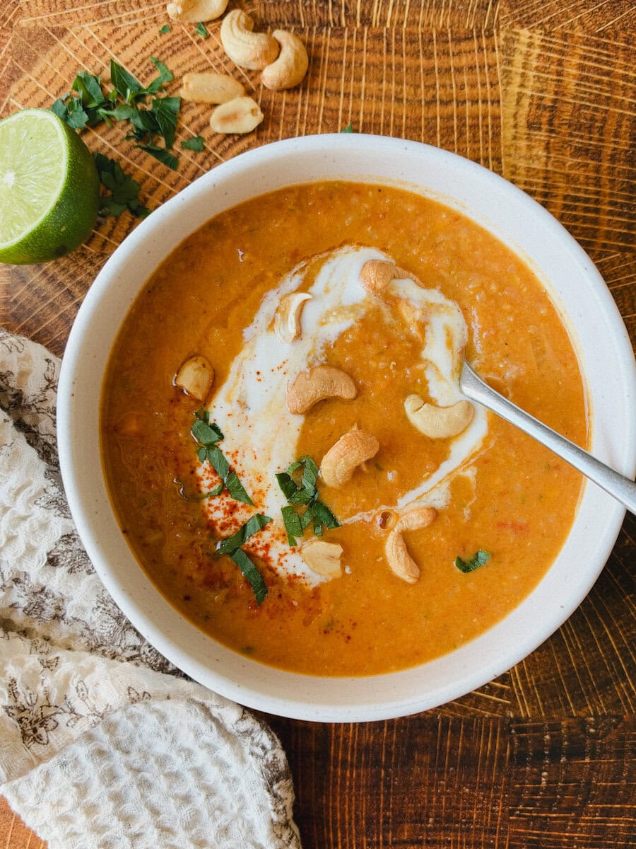 soupe aux lentilles corail dans un bol garni de yogourt, coriandre, noix de cajou sur table en bois avec linge et citron vert