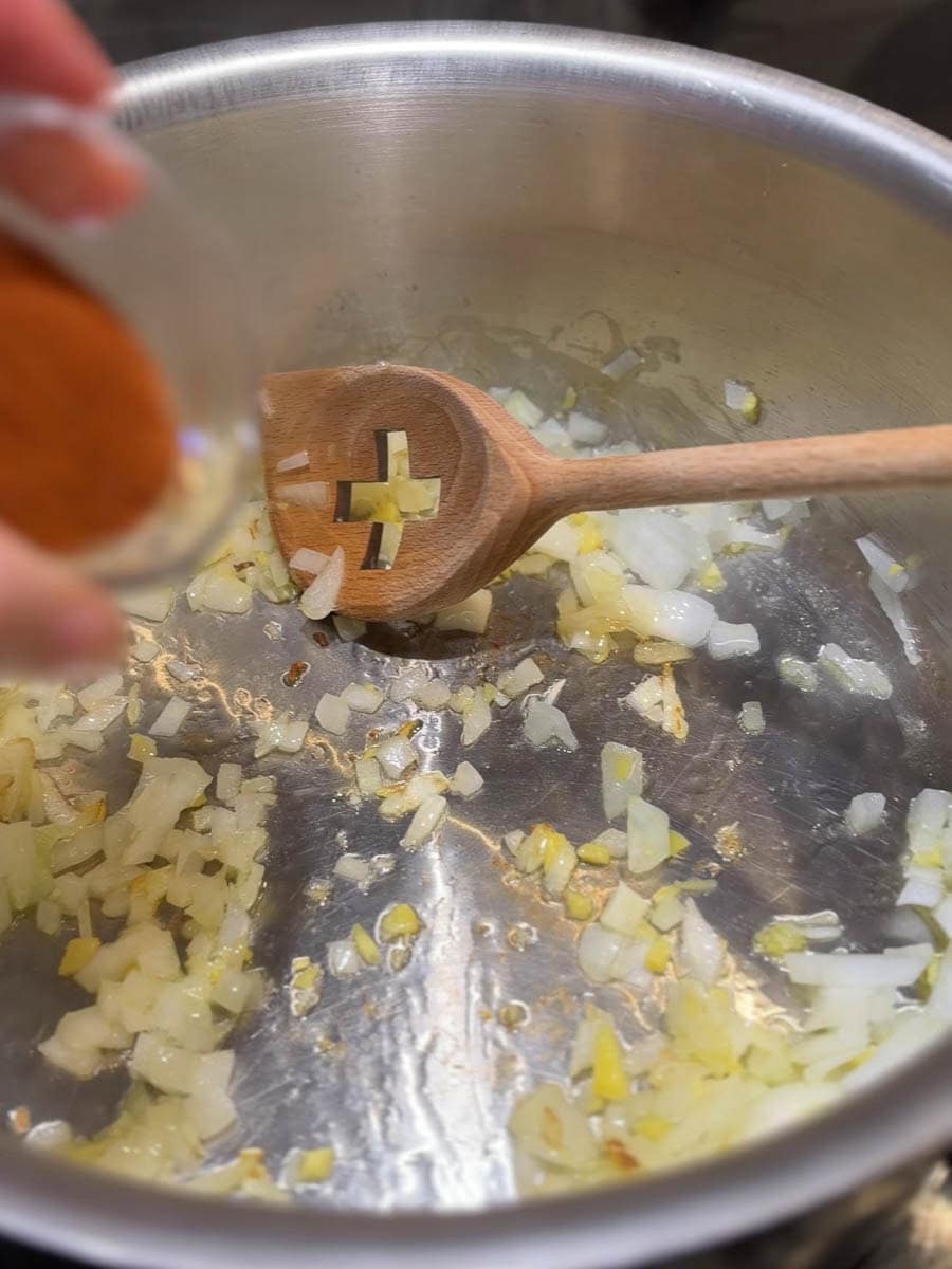 oignon et ail qui reviennent dans une casserole avec cuillère en bois et main qui verse les épices